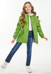 Žalia, vandeniui atspari striukė su gobtuvu, spaudžiamomis užtrauktukais, dviem priekiniais kišenėmis ir atspindinčiomis detalėmis; uždėta ant baltos marškinių ir mėlynų džinsų.