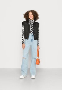 Colete puffer preto sobre um suéter de gola alta com padrão preto e branco, combinado com jeans rasgados azul claro e tênis brancos de plataforma, segurando uma bolsa laranja.
