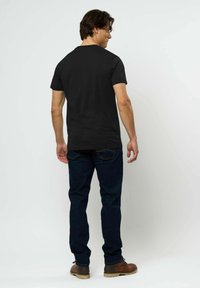 Vīrietis, kurš stāv ar muguru pret mums, valkājot vienkāršu melnu T-kreklu, tumši zilas džinsas un brūnas zābaki, pret vienkāršu baltu fonu.