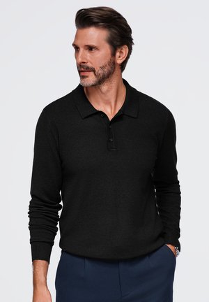 Un homme barbu portant un polo noir à manches longues et un pantalon bleu marine pose devant un fond blanc uni en regardant sur le côté.