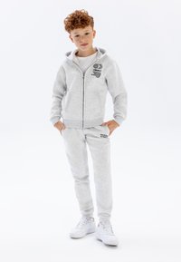 Lichtgrijze zip-up hoodie met voorzakken en zwarte grafische prints, gecombineerd met bijpassende joggingsbroek met tekst en elastische manchetten.
