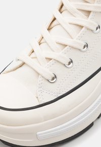 Basket en toile crème avec lacets plats, œillets argentés et semelle en caoutchouc noir. Dispose d'un bout arrondi et de coutures texturées.