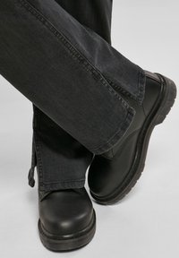 Persona que lleva botas de cuero negras y pantalones de pierna ancha gris oscuro con aberturas en los dobladillos, con los pies cruzados contra un fondo gris claro.