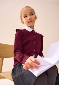 Bordeauxrode cardigan met ruches, witte kraag en knopen. Grijze geplooide rok. Kind zit, houdt witte bladzijden vast, met houten stoel op de achtergrond.