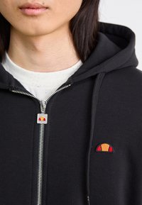 Czarna bluza z zamkiem błyskawicznym, z metalicznym suwakiem i wielokolorowym haftowanym logo na piersi, noszona na białej koszulce w kropki.