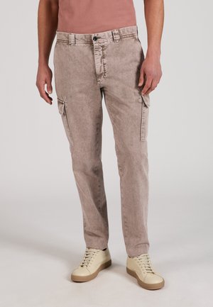 Mann trägt ausgebleichte beige Cargohosen mit Seitentaschen und cremefarbige Sneakers, steht vor schlichtem Hintergrund.