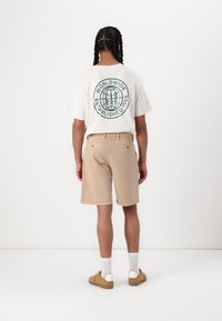 Camiseta blanca con gráfico de un globo azul en la parte posterior, combinada con pantalones cortos color beige. Calzado marrón con acentos blancos y calcetines cortos blancos. Fondo plano.