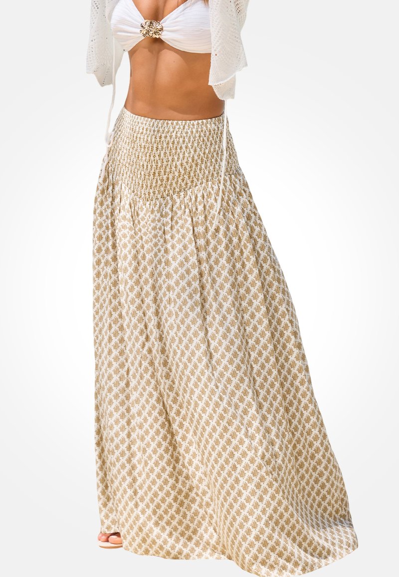 Vrouw draagt een lange beige rok met kleine bladprint, gecombineerd met een wit bikinitopje en een korte gebreide cardigan, gefotografeerd tegen een witte achtergrond.