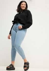 Camisa negra de botones con un nudo en la cintura, jeans de patrón azul claro y sandalias de plataforma negras. El conjunto tiene un corte relajado y un estilo casual.