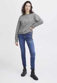 Sudadera gris con un diseño gráfico circular, combinada con jeans ajustados azules y botas de tobillo negras con un detalle de hebilla.