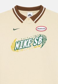 Polo crema con colletto a righe marroni, caratterizzato da una grafica verde e gialla con la scritta "NIKE SB" e un'etichetta rossa "Skateboarding".