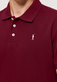 Bordový polo tričko vyrobené z texturované tkaniny, s klasickým límečkem, třemi knoflíky a malým bílým logem golfisty na hrudi.