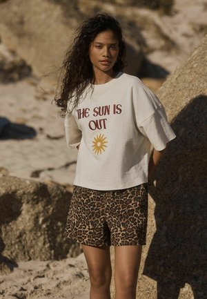 Kvinde med krøllet hår står på en klippefyldt strand iført en hvid skjorte med teksten "THE SUN IS OUT" og en sol-grafik, kombineret med leopardprint shorts.