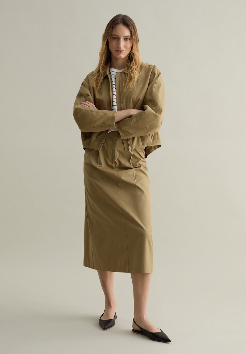 Giacca khaki cropped con maniche ampie sopra una gonna abbinata fino al ginocchio. La modella indossa delle ballerine nere a punta e una maglietta a righe bianche.