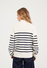 Kobieta z falowanymi włosami, ubrana w biało-granatowy sweter w paski, ciemne dżinsy i złote kolczyki w kształcie kół, zwrócona w lewo na jednolitym tle.