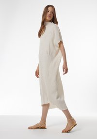 Witte jurk met een gestructureerd bladenpatroon, middellang, losse pasvorm. Gepaard met gouden platte sandalen met gekruiste banden.