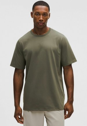 Giovane uomo in piedi, rivolto verso l'obiettivo, indossa una t-shirt a maniche corte verde oliva e pantaloni chiari su uno sfondo uniforme.