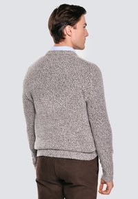 Muž otočený zády, oblečený v texturovaném šedém pleteném svetru přes světle modrou košili s límečkem a v hnědých kalhotách, stojící proti jednobarevnému pozadí.