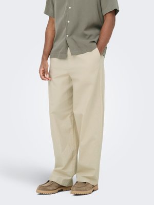 Homme portant un pantalon beige ample, des chaussures en daim marron clair et une chemise à manches courtes vert olive boutonnée, debout avec une main dans la poche.