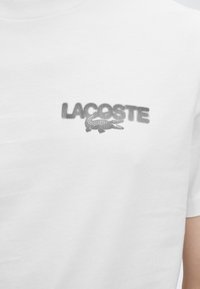 Λευκό T-shirt με υφασμάτινο μαύρο λογότυπο Lacoste και το έμβλημα αλιγάτορα στο κέντρο του στήθους.