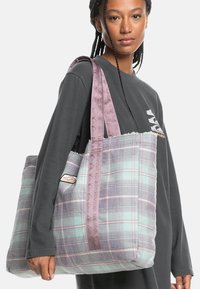 Karierte Tragetasche in Pastellgrün und Lila. Weicher Stoff, gepolsterte Seiten und ein lila Riemen mit eingeprägtem Logo-Detail.
