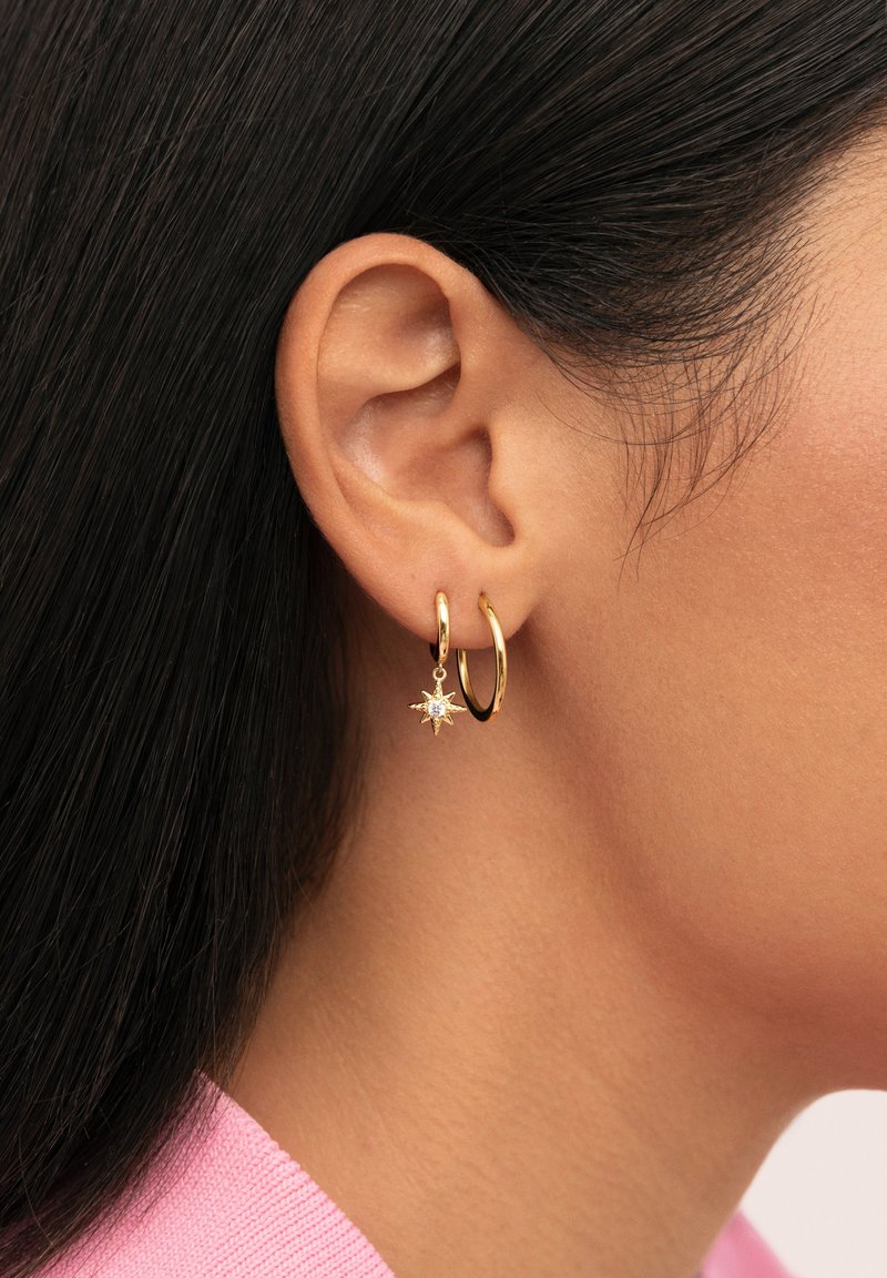 Pendientes de aro de oro con un charm en forma de estrella, que incluye una piedra preciosa clara. El diseño es elegante y moderno, sobre un fondo de cabello oscuro.