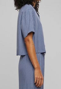 Camisa de manga corta azul con rayas verticales blancas, con cuello y diseño recortado. Tejido suave con un ajuste holgado.