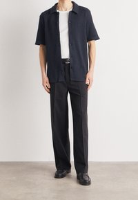 Marineblaues kurzärmeliges Hemd mit Knöpfen und einem gerippten Muster, darunter ein weißes T-Shirt mit Rundhalsausschnitt, kombiniert mit schwarzen Hosen und schwarzen Schuhen.