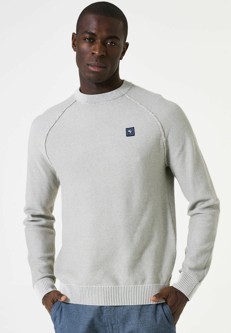 Lichtgrijze gebreide trui met een ronde hals, geribbelde mouwen en onderrand. Heeft een klein blauw logo-opdruk op de borst. Gladde textuur.