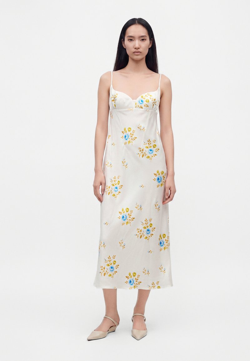 Femme portant une robe nuisette midi blanche avec des motifs floraux bleus et jaunes et des ballerines pointues beiges, debout devant un fond blanc uni.