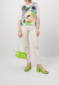 Bunt gemusterte Bluse aus weichem Stoff; beige, enganliegende Hose; grüne Plateausandalen; kleine grüne Umhängetasche mit Kordelzug.