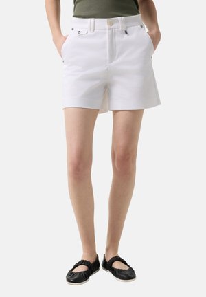 Vrouw draagt witte high-waist shorts met voorzakken en zwarte platte schoenen, handen in de zakken, staand tegen een effen achtergrond.