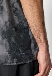 Mandearm med en lille rød tatovering ved siden af en sort og grå mønstret Climacool skjorte og sorte shorts mod en ensfarvet baggrund.