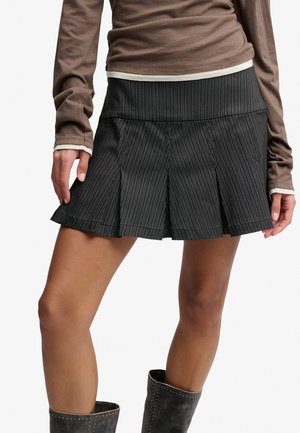 Kobieta nosząca czarną plisowaną mini spódniczkę w drobną prążek, brązowy longsleeve i czarne buty na białym tle.