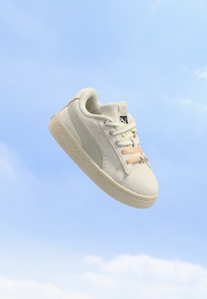 Baskets blanches Puma avec semelle crème et clips élastiques beiges pour lacets flottant sur un fond de ciel bleu.