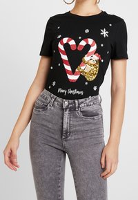 Svart kortärmad t-shirt med en glittrig guldslo, röd-vita randiga polkagrisar, snöflingor och texten "God Jul".