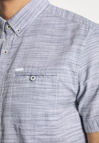 Camisa de botones de manga corta en azul claro con un patrón texturizado. Presenta un bolsillo en el pecho con botón y un diseño de cuello casual.