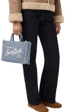 Personne portant un jean noir, des chaussures marron et une veste en shearling beige, tenant un sac en velours côtelé bleu avec le texte "Rêver de Saint Barth".