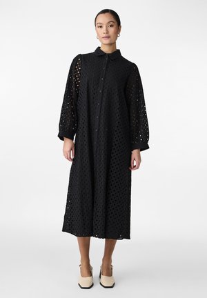 Schwarzes Hemdkleid mit durchgehender Knopfleiste, strukturiertem Stoff mit offenen Ausschnitten, langen Ärmeln und Kragen. Kombiniert mit cremefarbenen Blockabsatzschuhen.