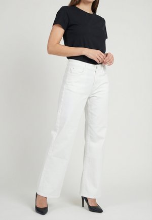 Donna con maglietta nera a maniche corte, jeans bianchi a vita alta e gamba larga, e scarpe nere con tacchi alti, in piedi contro uno sfondo neutro.