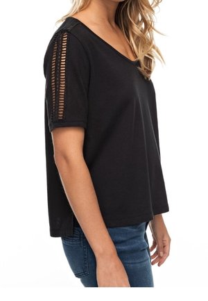 Femme portant un haut noir à manches courtes avec des détails en maille ouverte sur les manches, assorti à un jean bleu, vue de profil.