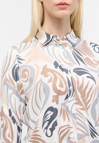 Donna che indossa una camicetta con bottoni con motivi astratti di foglie e vortici beige, grigi e neri su uno sfondo bianco.