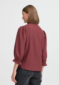 Rostfarbene Bluse mit strukturiertem Finish, gepufften Ärmeln und Rüschensäumen. Die Rückansicht hebt eine entspannte Passform und dezente Designdetails hervor.