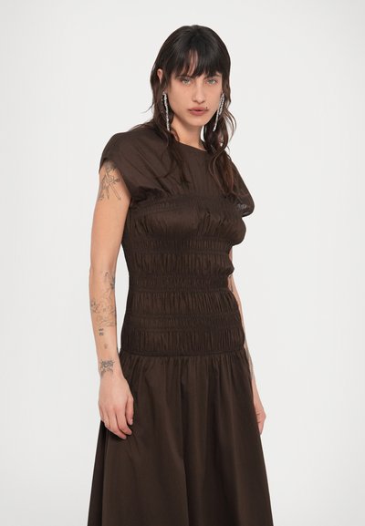 Νεαρό άτομο με μακριά σκούρα μαλλιά, ντυμένο με ένα σκούρο καφέ φορεματάκι με πιέτες και μακριά κρεμαστά σκουλαρίκια, στέκεται μπροστά σε απλό λευκό φόντο.