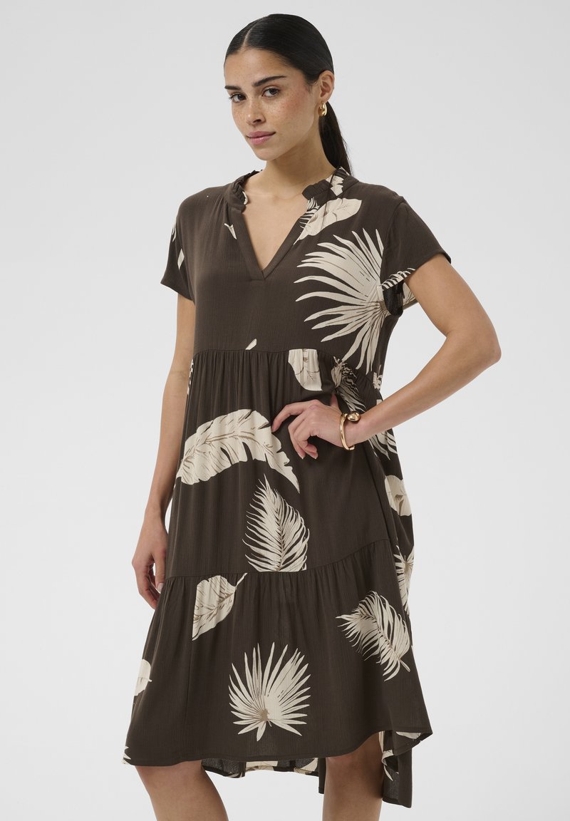 Femme aux cheveux foncés portant une robe ample marron foncé avec des imprimés de feuilles tropicales blanches et des manches courtes, posant avec une main sur la hanche.