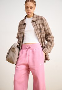 Bruin geruit flanellen shirt over een witte tanktop, in combinatie met roze sweatpants met een witte Nike-logo, en een grijze schoudertas.