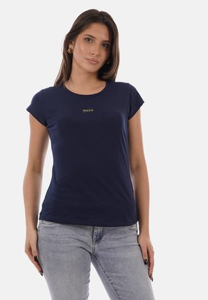 Donna che indossa una maglietta aderente blu navy con il logo "YES-ZEE" e jeans grigio chiaro, in piedi davanti a uno sfondo chiaro e semplice.