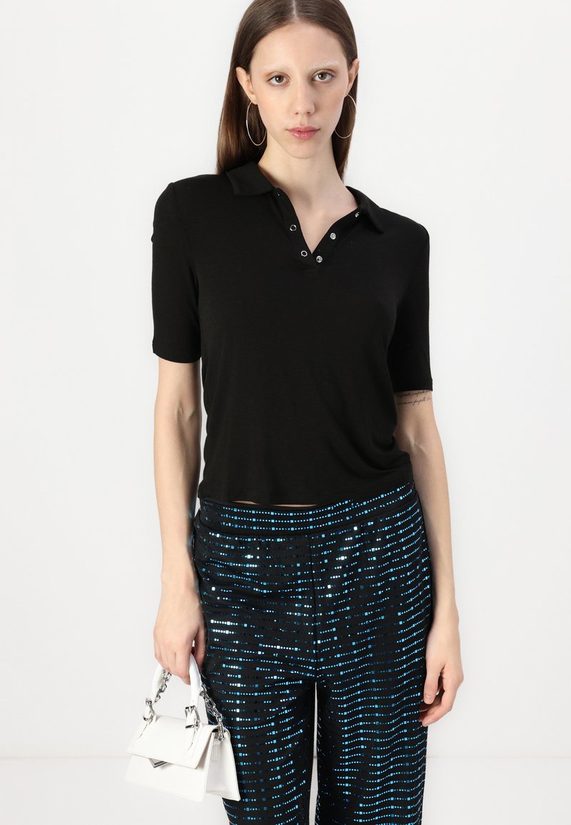 Polo nero con maniche corte e colletto abbottonato, abbinato a pantaloni neri con strisce metalliche blu. Tiene una piccola borsa bianca.