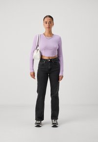 Lilac cropped long-sleeve top, black jeans, and sneakers. Model holds a small white bag. Simple design with ribbed texture and chain accessory.