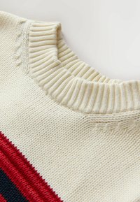 Kremowy sweter w dzianinę z teksturowanym ściągaczem, z czerwonymi i granatowymi poziomymi paskami na całym ciele. Miękki, średniej wagi materiał.